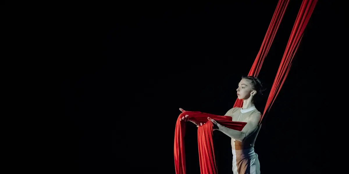 Arielle Gorodetskaya, une artiste aérienne vêtue d'un costume blanc et beige, tient de longs rubans de soie rouge alors qu'elle se prépare pour son numéro aérien captivant sur un fond sombre.