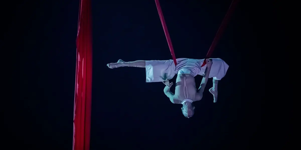Arielle Gorodetskaya, une artiste aérienne, est suspendue à l'envers dans les airs, en équilibre sur des soies rouges, les bras tendus et les jambes allongées, vêtue d'un costume blanc sur un fond sombre.