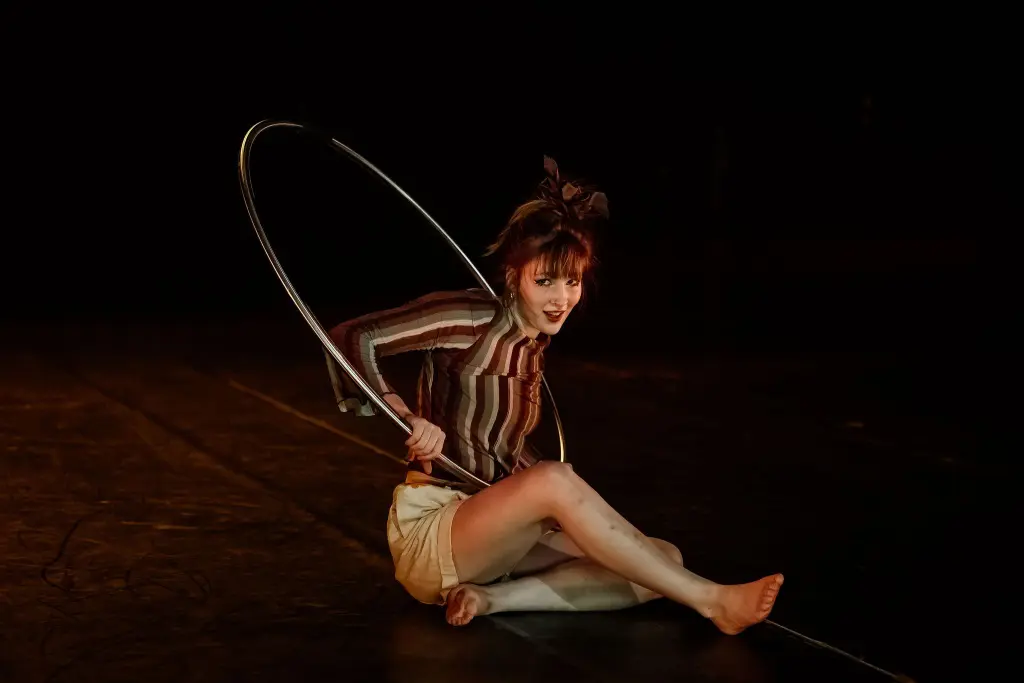 Victoria Baudry-McPherson, coiffée d'un chignon, est assise, souriante, sur une scène faiblement éclairée, tenant un grand cerceau métallique autour de ses épaules et de ses genoux. Elle porte un haut rayé et un short de couleur claire.