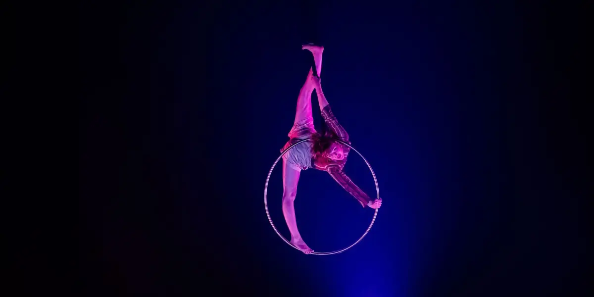 Une artiste aérienne, Victoria Baudry-McPherson, en éclairage rose, est suspendue à l'envers à un cerceau, étendant gracieusement une jambe vers le haut sur un fond bleu foncé et violet.