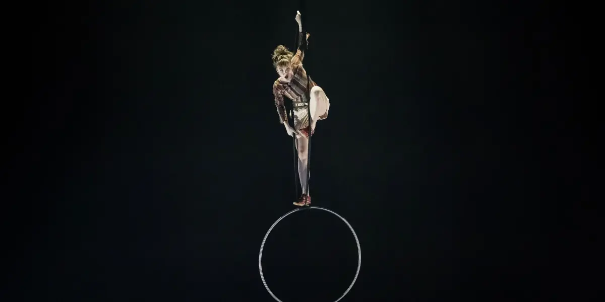 L'artiste aérienne Victoria Baudry-McPherson se tient en équilibre sur une jambe au sommet d'un cerceau suspendu sur un fond sombre, saisissant le cerceau d'une main et tendant l'autre vers le haut en direction du crochet.