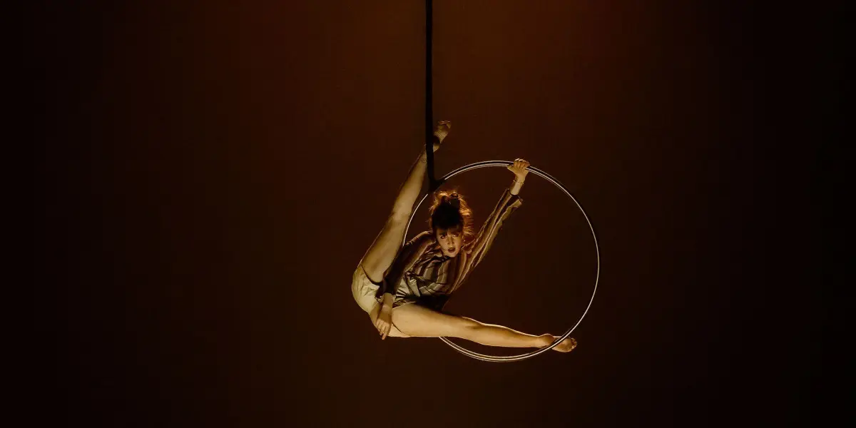 Victoria Baudry-McPherson se produit sur un cerceau aérien, suspendue dans les airs, une jambe tendue vers le haut et l'autre vers l'extérieur, saisissant le cerceau d'une main, sur un fond sombre éclairé par des projecteurs.