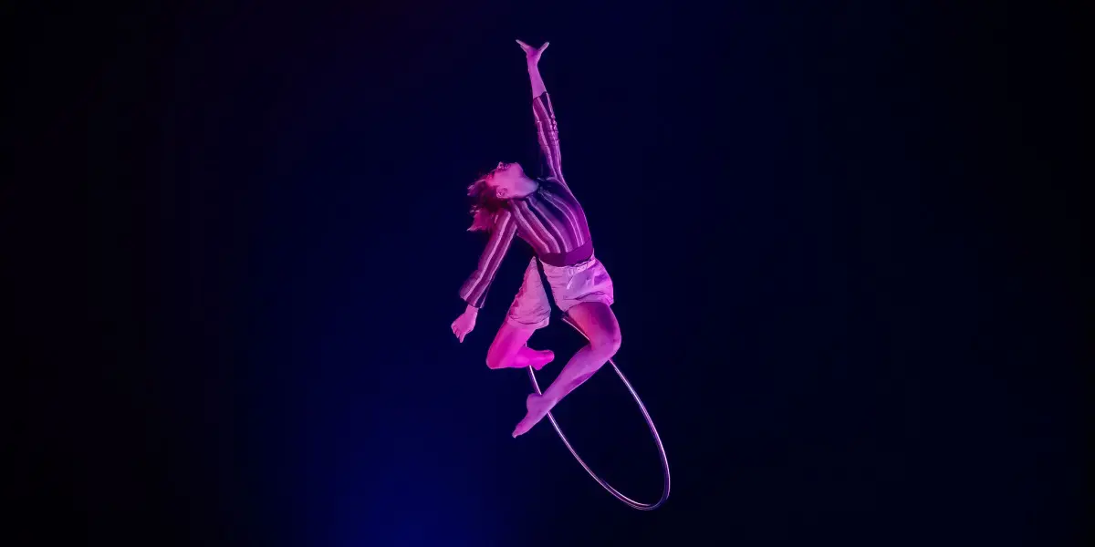 L'artiste aérienne Victoria Baudry-McPherson, vêtue d'une chemise et d'un short rayés, se suspend gracieusement à un cerceau, un bras tendu vers le haut, sur un fond sombre éclairé par des lumières roses et bleues spectaculaires.