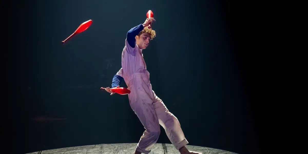 Un artiste pieds nus, Merlin Matthewson, vêtu d'une combinaison blanche et d'une chemise bleue, jongle avec trois épingles rouges sur une scène faiblement éclairée, deux épingles en l'air et une dans la main. Un éclairage dramatique met en valeur l'artiste.