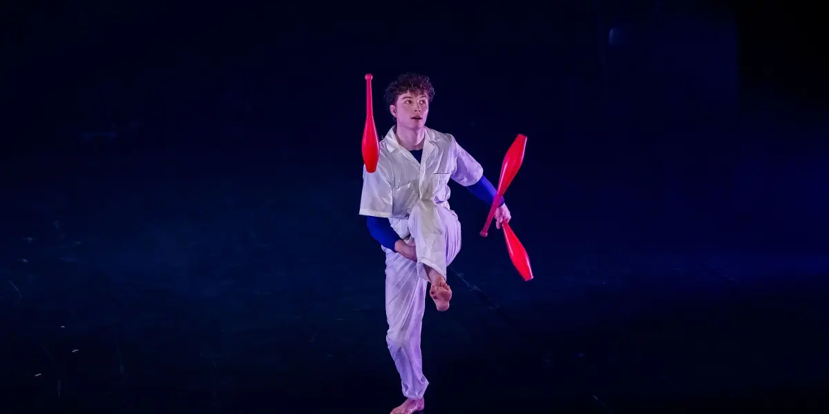 Merlin Matthewson, vêtu de blanc, fait un numéro de jonglerie sur scène, se tenant en équilibre sur une jambe et lançant deux massues rouges en l'air sur un fond sombre éclairé par des projecteurs.