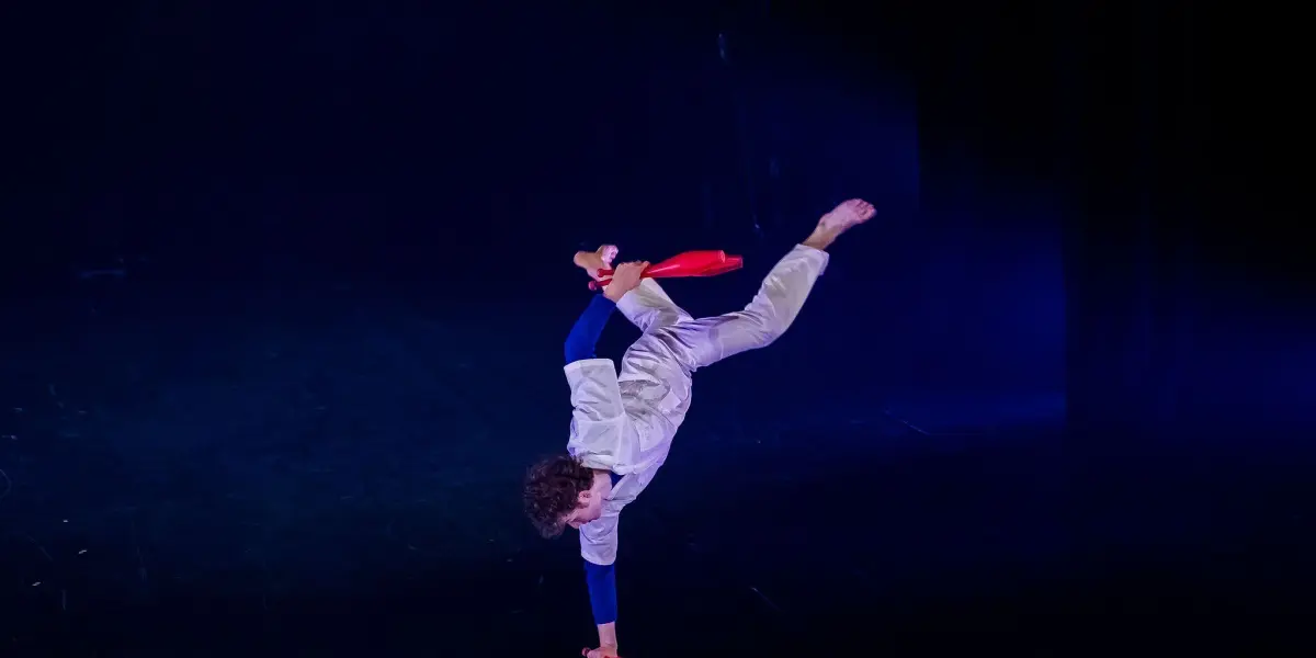 Un artiste, Merlin Matthewson, se tient en équilibre sur une main sur une scène sombre, vêtu d'une tenue blanche et tenant deux massues de jonglage rouges, avec une jambe pliée et l'autre tendue en l'air.