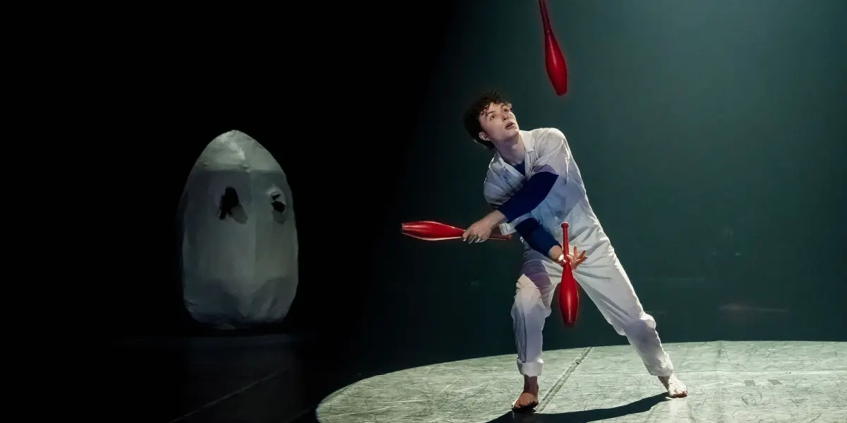 Un artiste en tenue blanche, peut-être Merlin Matthewson, jongle avec des massues rouges sous un projecteur sur scène. À l'arrière-plan, un grand objet blanc de forme ovale avec des marques noires est visible dans l'ombre.