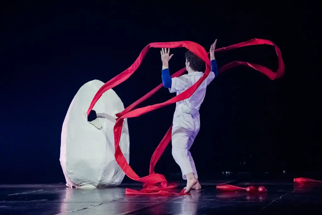 Un artiste en blanc, peut-être Merlin Matthewson, tient des rubans rouges sur scène près d'une grande structure abstraite blanche avec un trou, sur un fond sombre.