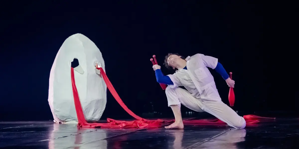 Un artiste en costume blanc prend une pose théâtrale sur scène, tenant des massues de jonglage rouges, avec des rubans rouges reliés à un grand accessoire ovale blanc derrière lui. La scène théâtrale, conçue par Merlin Matthewson, est éclairée de manière saisissante sur une toile de fond sombre.
