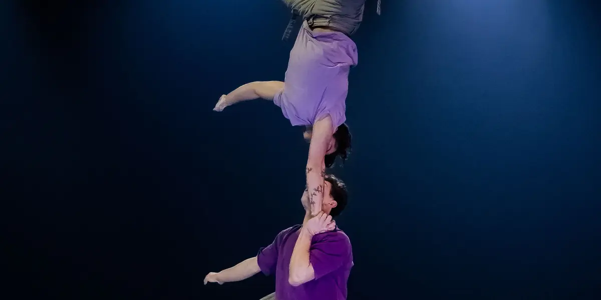 Deux acrobates se produisent sur scène ; l'un se tient en équilibre sur la main de l'autre dans un poirier à un bras. En tenue décontractée et sous un éclairage dramatique, ils font preuve d'une remarquable habileté, rappelant les célèbres performances de Frédéric Langevin.