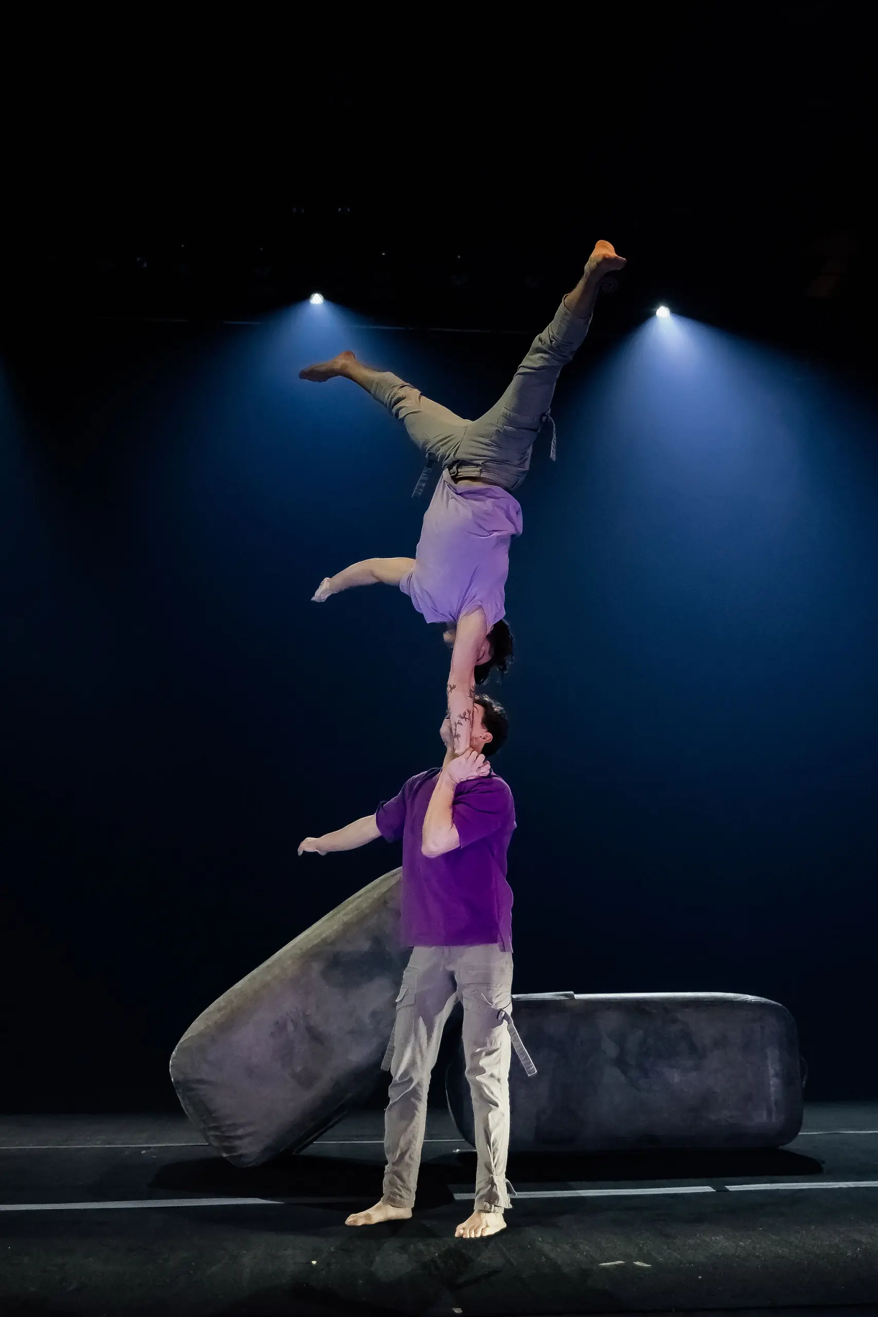 Deux acrobates se produisent sur scène ; l'un se tient en équilibre sur la main de l'autre dans un poirier à un bras. En tenue décontractée et sous un éclairage dramatique, ils font preuve d'une remarquable habileté, rappelant les célèbres performances de Frédéric Langevin.