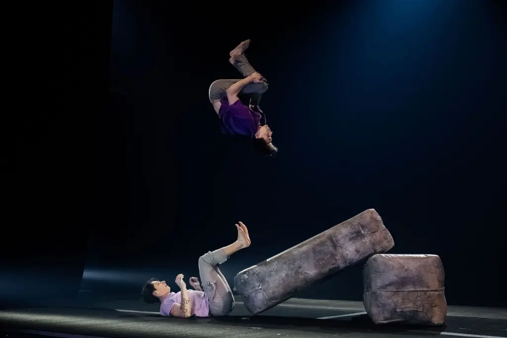 Deux artistes en tenue décontractée font des acrobaties sur scène ; l'un est allongé sur le sol, les jambes levées, tandis que Frédéric Langevin se balance au-dessus, dans les airs, avec de gros blocs rembourrés à proximité, sous un éclairage dramatique.
