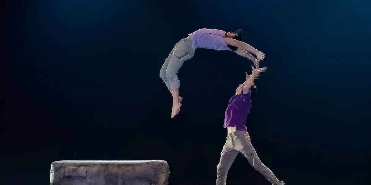 Deux acrobates se produisent sur scène ; une personne saute en arrière en plein vol tandis que Frédéric Langevin, debout, tend la main pour la rattraper. Un bloc rectangulaire rembourré est placé à proximité, sous les lumières de la scène.