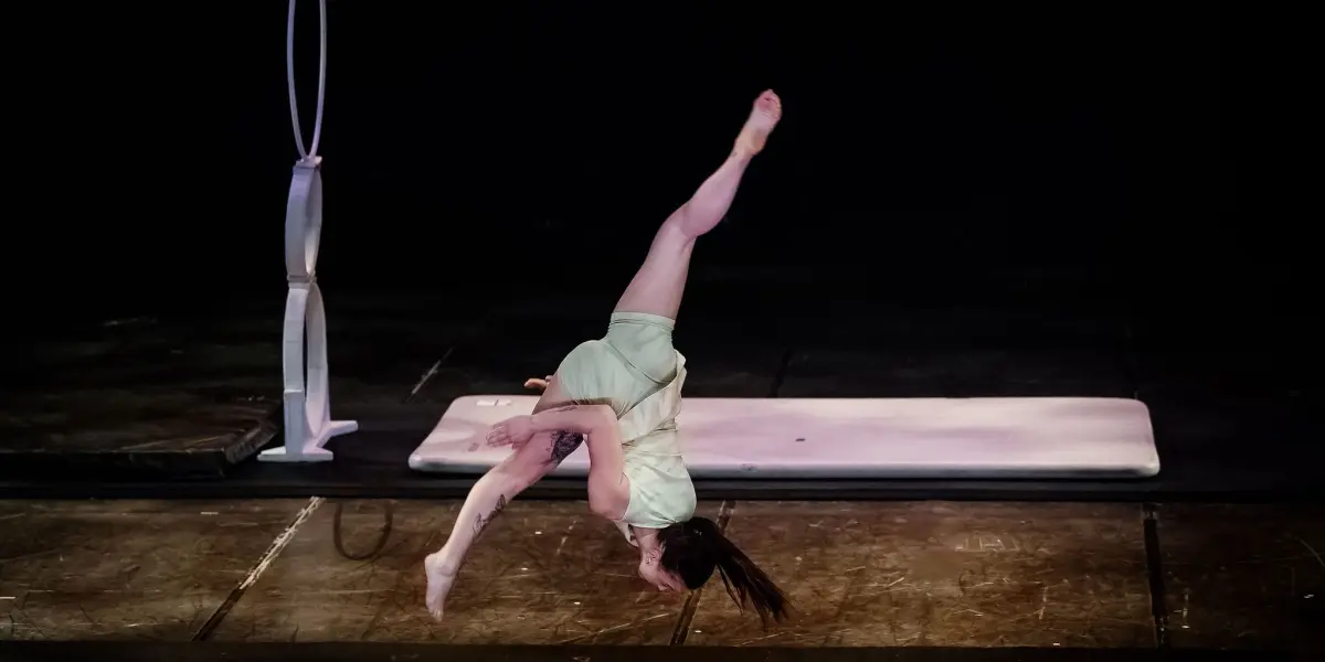 Une gymnaste en tenue vert clair effectue une roue à une main sur la scène, avec une jambe tendue vers le haut. Le matériel de gymnastique Chanel Monange et un tapis sont visibles dans l'arrière-plan faiblement éclairé.