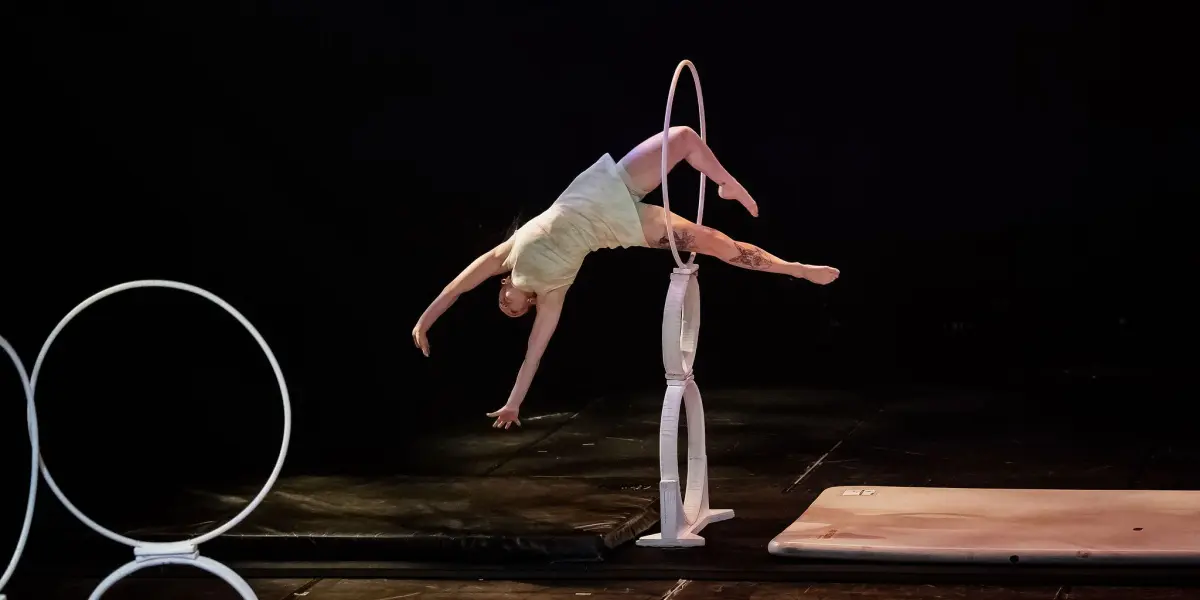 Une artiste de cirque, Chanel Monange, se tient gracieusement en équilibre sur une pile d'anneaux blancs, une jambe étendue et les bras tendus, sur un fond de scène sombre. D'autres anneaux et tapis sont visibles à proximité.