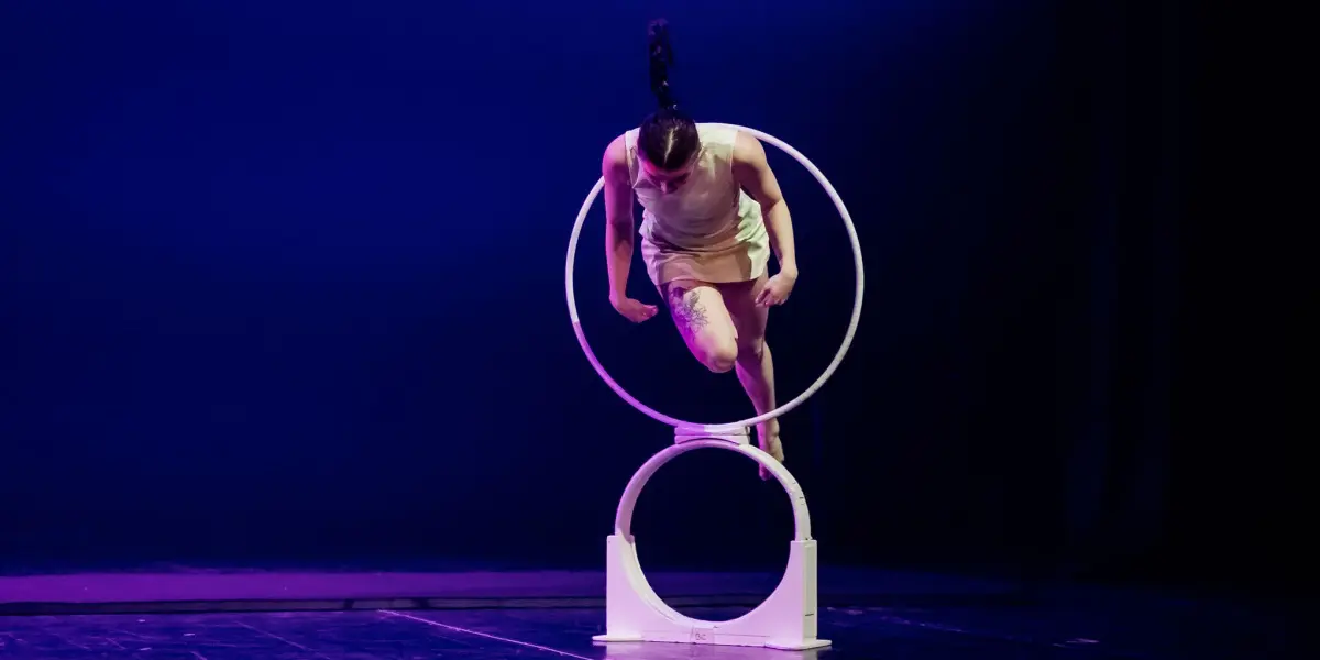 Chanel Monange, avec une longue queue de cheval et une tenue de couleur claire, saute acrobatiquement à travers deux cerceaux blancs empilés sur une scène à l'éclairage violet dramatique.
