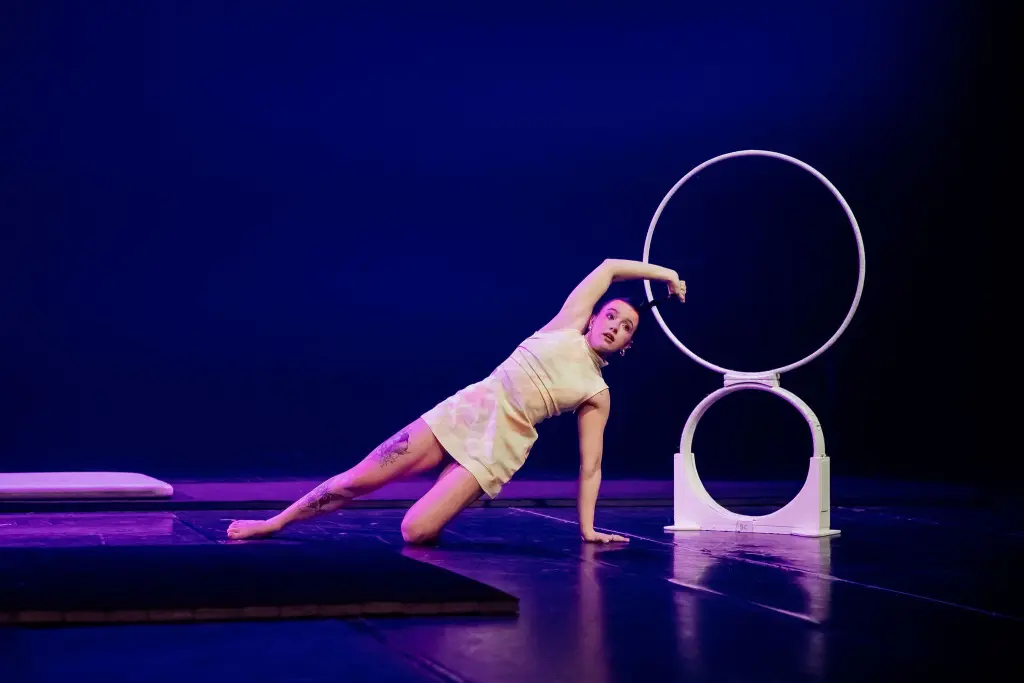 Une artiste en tenue claire se tient en équilibre sur un bras à côté d'arceaux circulaires blancs sur une scène sombre, avec un éclairage violet dramatique soulignant la scène - un moment élégant de Chanel Monange.