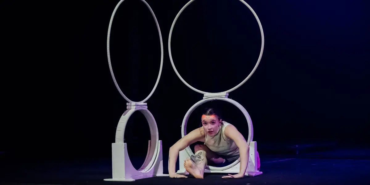 Chanel Monange, une contorsionniste talentueuse, se produit sur scène, rampant dans un anneau circulaire blanc posé sur le sol, avec deux anneaux verticaux plus grands derrière elle, sous un éclairage dramatique.