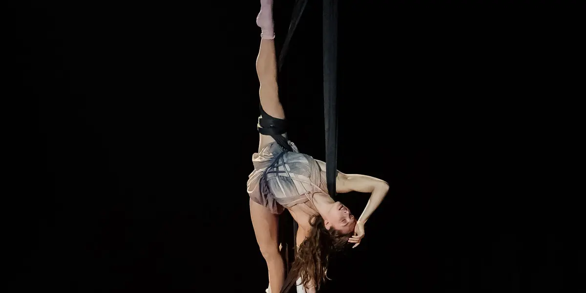 L'artiste aérienne Rose Munger, vêtue d'une tenue légère, est suspendue la tête en bas à des soies aériennes noires, posant gracieusement avec une jambe tendue et un bras reposant sur son front, sur un fond sombre.