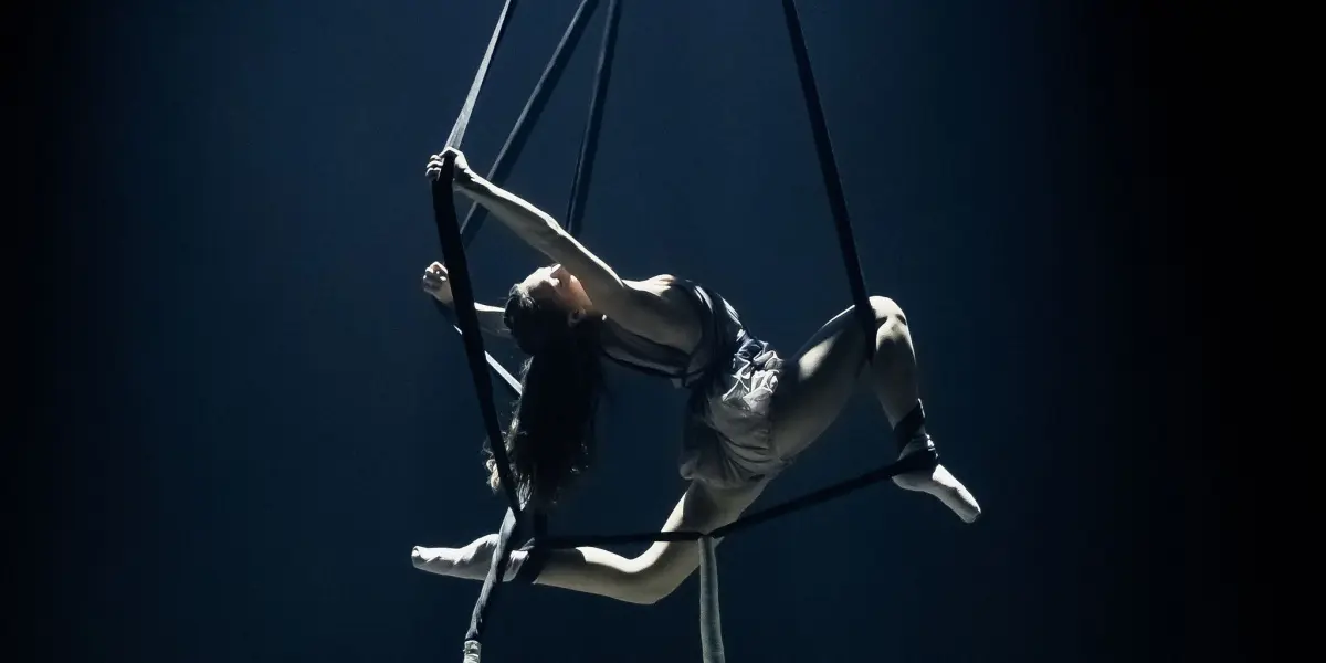 Une voltigeuse, Rose Munger, exécute une pose acrobatique sur un trapèze suspendu, éclairée par un projecteur sur un fond sombre. Elle étire ses membres avec grâce, mettant en valeur sa force et sa souplesse.