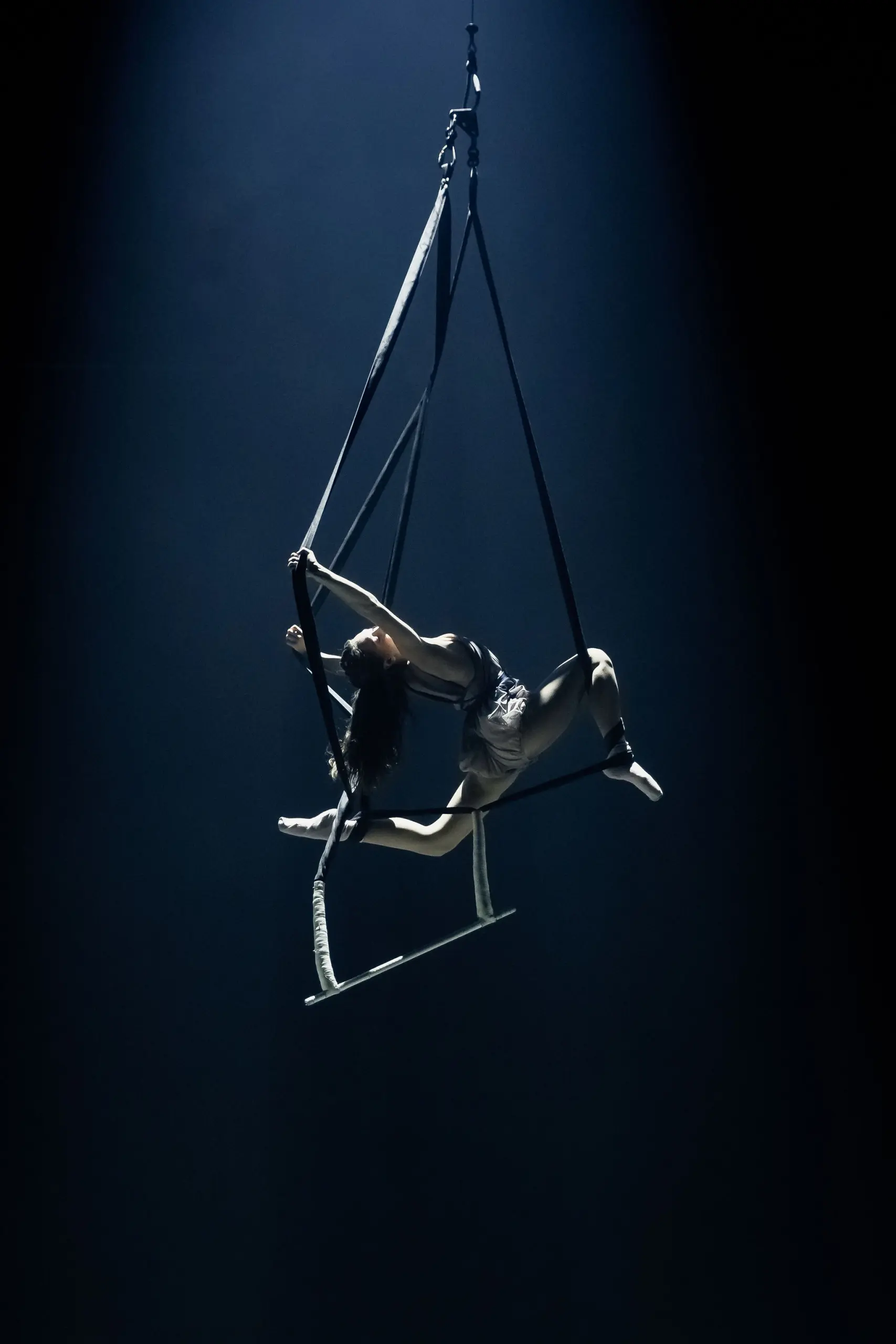 Une voltigeuse, Rose Munger, exécute une pose acrobatique sur un trapèze suspendu, éclairée par un projecteur sur un fond sombre. Elle étire ses membres avec grâce, mettant en valeur sa force et sa souplesse.