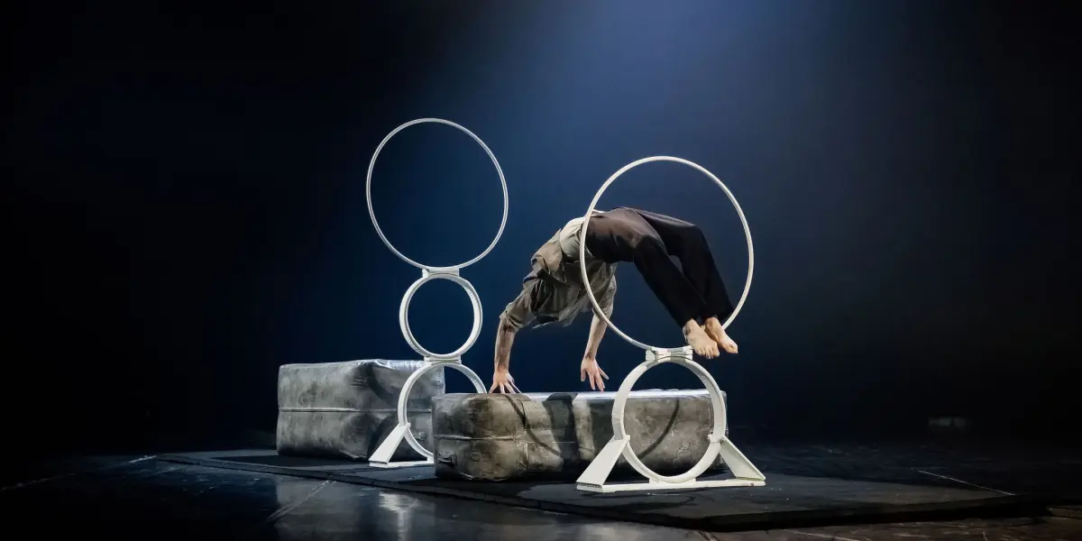 Un interprète, inspiré par Frédéric Langevin, se tient en équilibre sur les mains, les jambes étendues à travers un grand anneau métallique, placé sur des blocs rembourrés, sous un éclairage de scène dramatique dans un théâtre sombre.
