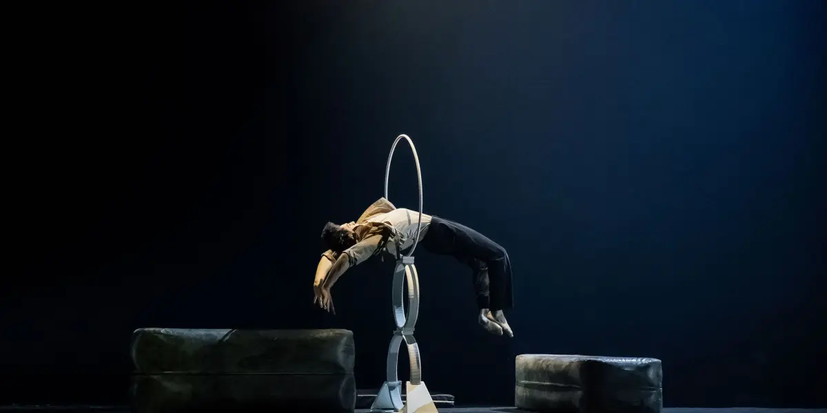 Frédéric Langevin se tient en équilibre horizontal dans un cerceau circulaire, soutenu par une structure, les bras ballants et les jambes étendues. Deux grands coussins encadrent la scène, tandis que la scène faiblement éclairée s'estompe pour laisser place à un arrière-plan sombre.