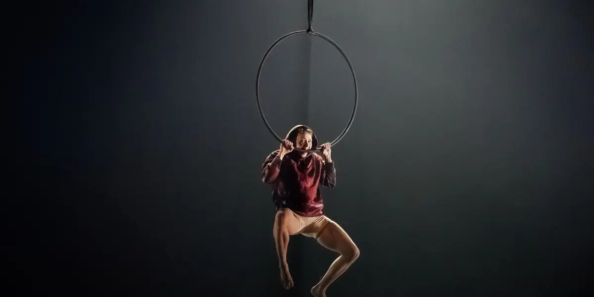 Sarah Norden, vêtue d'un haut marron et d'un short beige, se suspend gracieusement à un cerceau aérien, éclairé par un projecteur sur un fond sombre.
