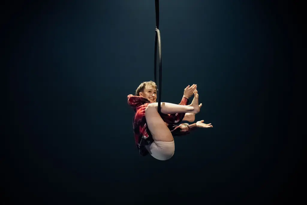 L'artiste aérienne Sarah Norden, vêtue d'une chemise à carreaux rouge et d'un short clair, est suspendue à l'envers dans un cerceau, souriant, les jambes pliées et se tenant aux pieds, sur un fond sombre.