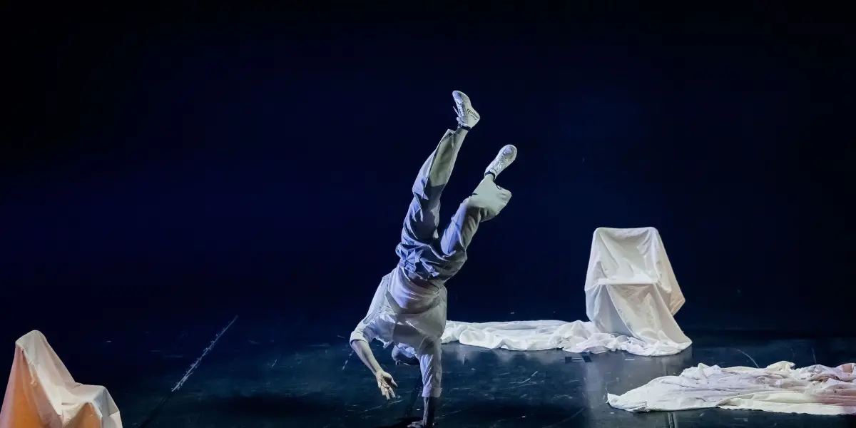 Un danseur vêtu de blanc, peut-être Matthieu Larose, effectue un équilibre sur une main sur scène, entouré de draps blancs drapant des objets, sur un fond sombre.