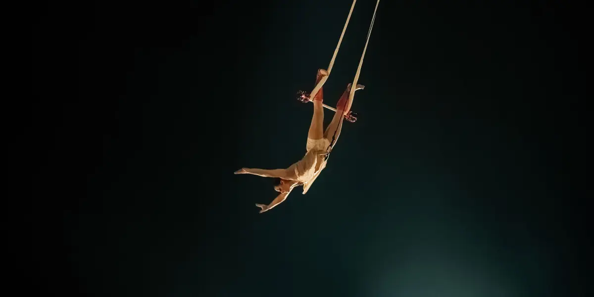 Une acrobate vêtue d'une tenue beige, rappelant l'élégance de Malou Latrompette, effectue une manœuvre aérienne sur des cordes suspendues sur un fond sombre, les bras et les jambes gracieusement tendus.