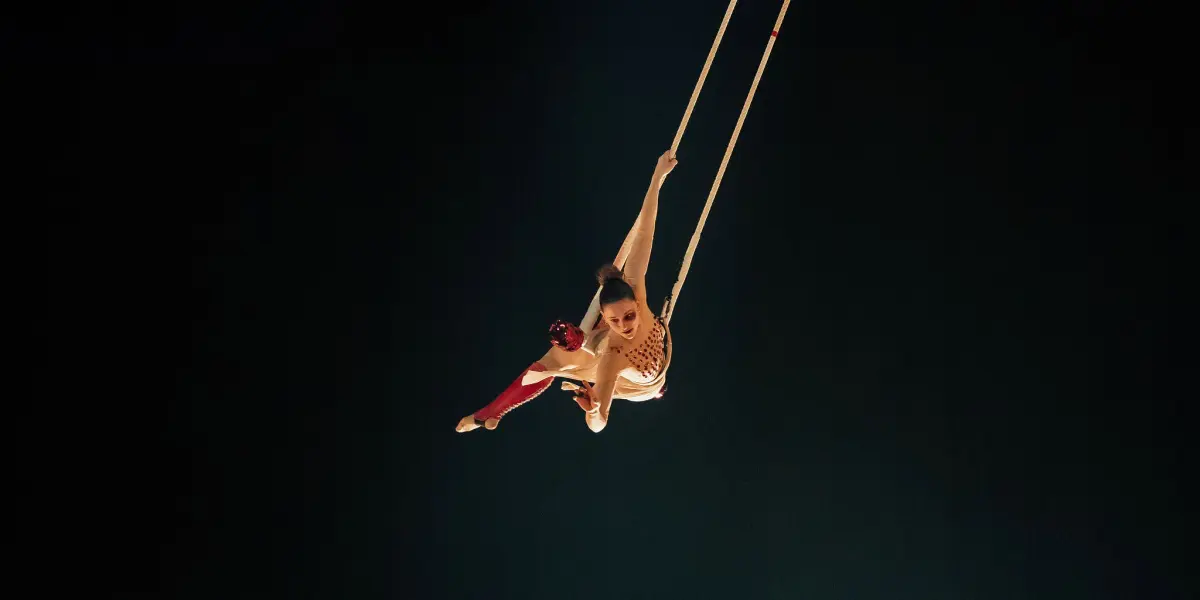 Malou Latrompette, une trapéziste vêtue d'un costume blanc et rouge, évolue en hauteur, suspendue par un bras à un trapèze, sur un fond sombre qui met en valeur sa pose dans les airs.