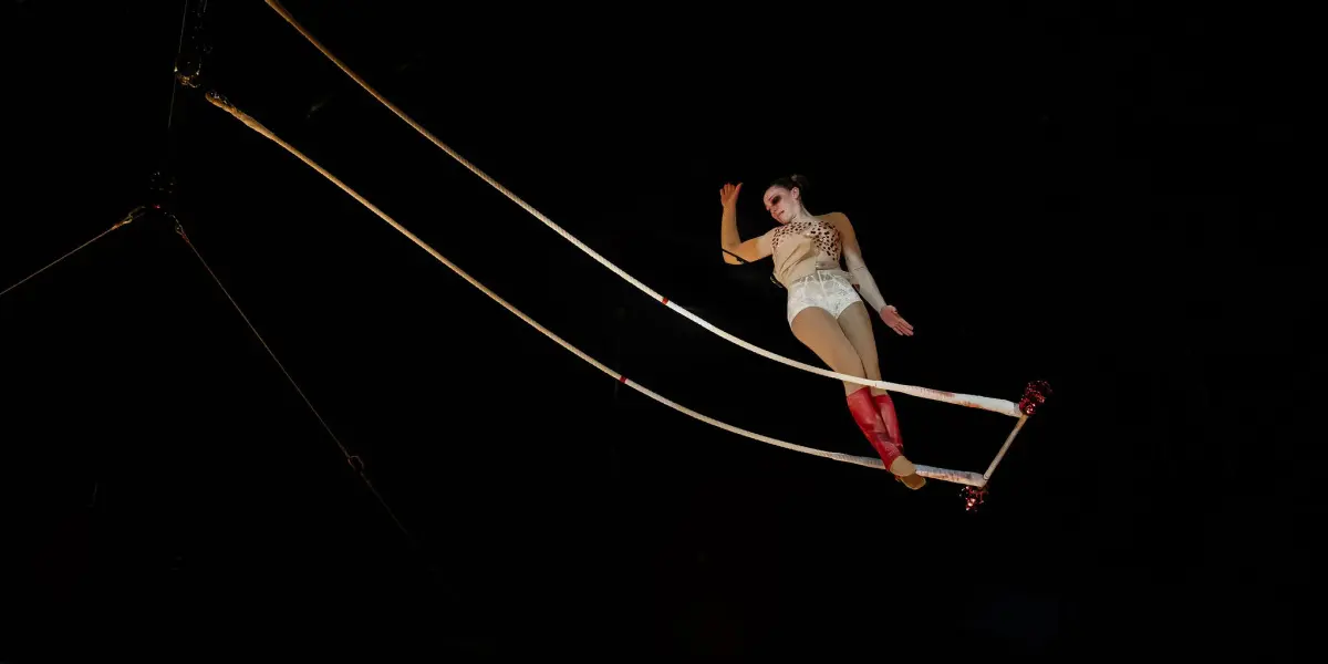 Malou Latrompette, une voltigeuse de cirque vêtue d'un costume blanc et rouge, se produit sur un trapèze suspendu, prenant une pose dramatique au-dessus du sol sur un fond sombre.