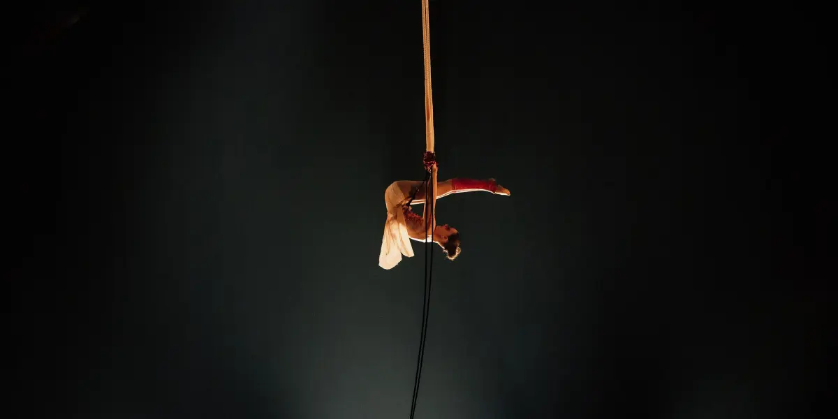 Un artiste de cirque, Malou Latrompette, vêtu d'un costume blanc, est suspendu à une corde en l'air, les jambes enroulées autour de la corde, sur un fond sombre éclairé par des projecteurs doux.