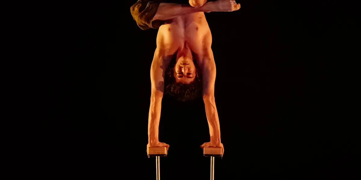 Thomas Parent, torse nu, se tient en équilibre sur deux cannes, les jambes pliées et croisées, sur une scène faiblement éclairée, parsemée de feuilles et de débris.