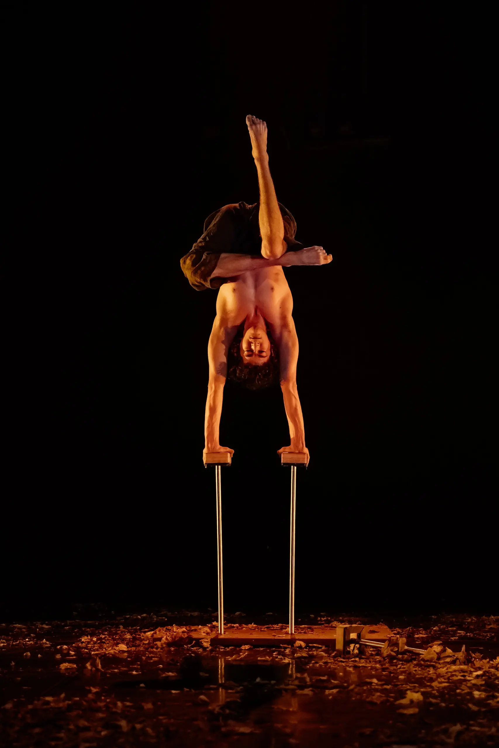 Thomas Parent, torse nu, se tient en équilibre sur deux cannes, les jambes pliées et croisées, sur une scène faiblement éclairée, parsemée de feuilles et de débris.