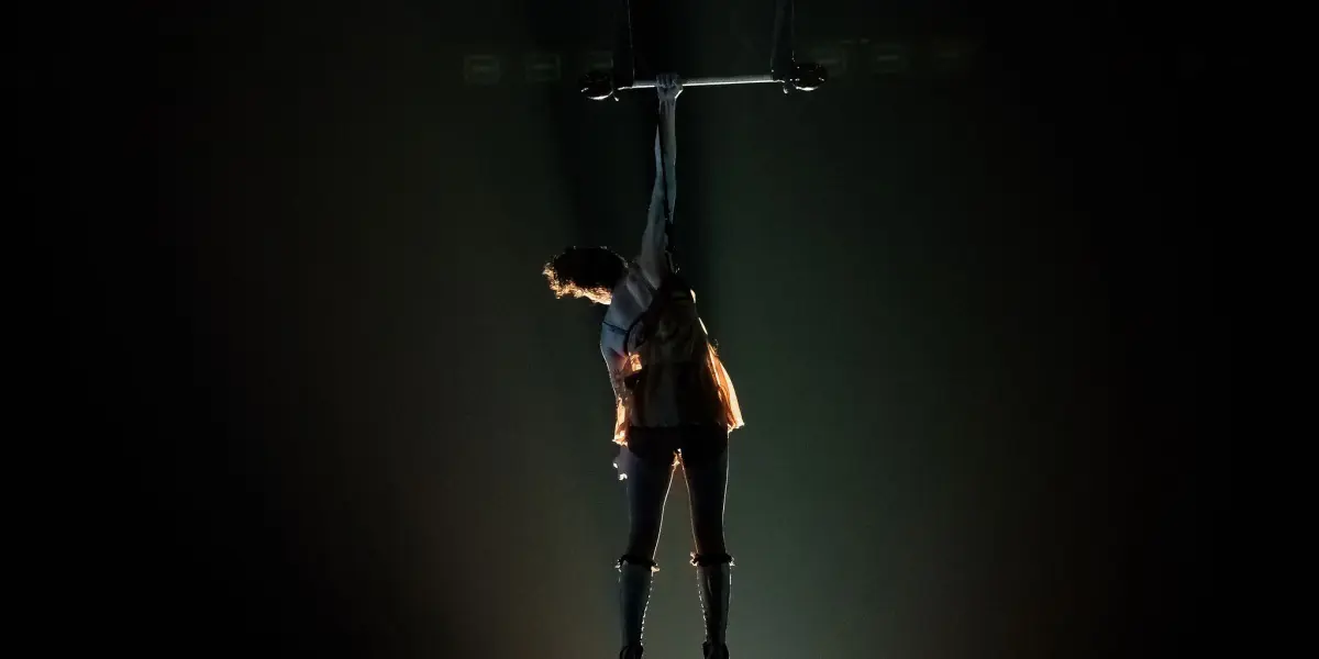 L'artiste aérienne Alizé Poitreau est suspendue par un bras à un trapèze au-dessus d'une scène sombre, éclairée par un projecteur dramatique, les jambes pointées vers le bas et le corps légèrement arqué.