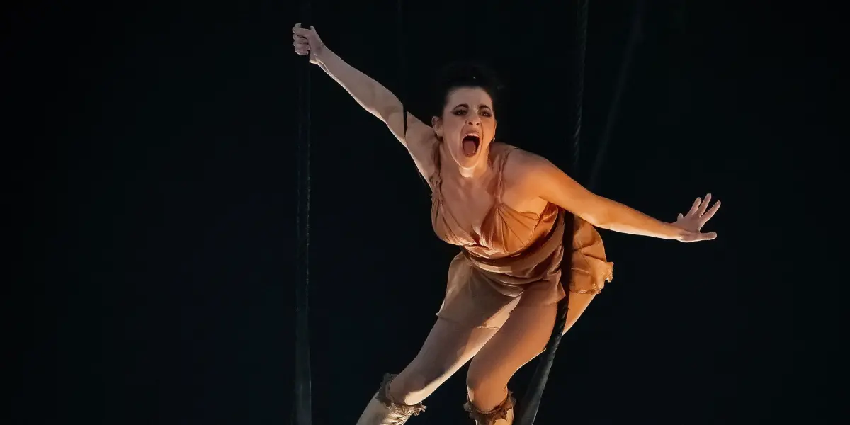 Alizé Poitreau, une trapéziste vêtue d'un costume beige et de bottes à hauteur des genoux, se balance dans les airs, une main agrippée à la corde, l'autre bras tendu, sur fond de lumières vives et d'obscurité.