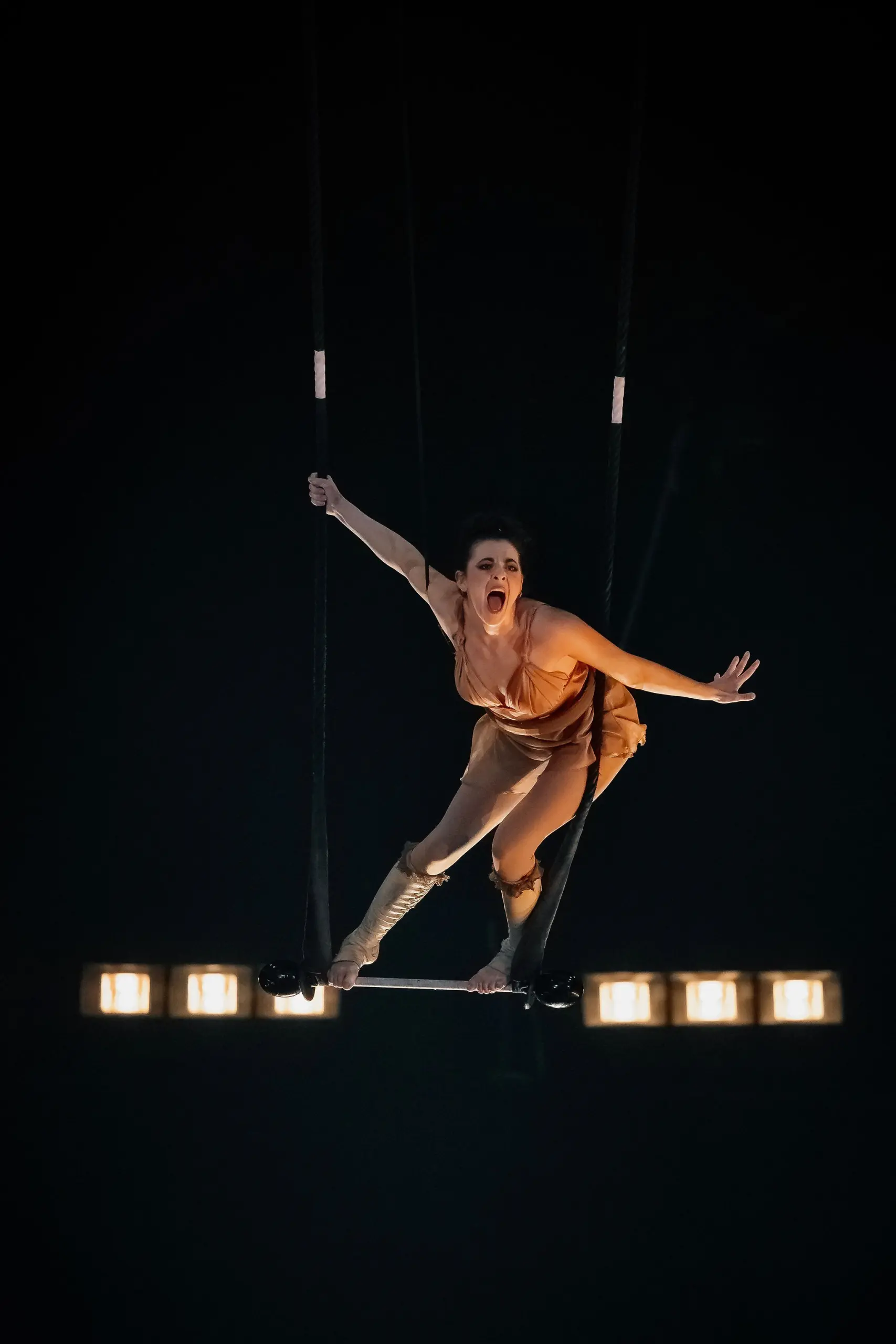 Alizé Poitreau, une trapéziste vêtue d'un costume beige et de bottes à hauteur des genoux, se balance dans les airs, une main agrippée à la corde, l'autre bras tendu, sur fond de lumières vives et d'obscurité.