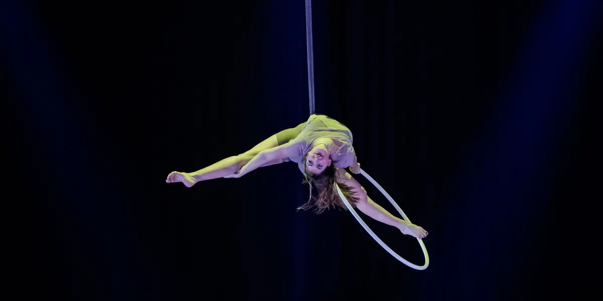 L'artiste aérienne Kristina Shelton, vêtue d'une tenue verte, est suspendue à l'envers à un cerceau, les jambes écartées, sous un éclairage de scène dramatique et un arrière-plan sombre.