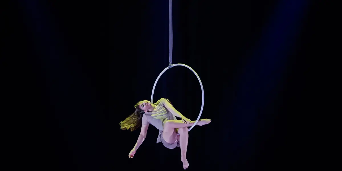 Kristina Shelton, vêtue d'un costume léger, évolue gracieusement sur un cerceau aérien suspendu sur un fond sombre, les lumières de la scène la mettant en valeur et créant une atmosphère dramatique.
