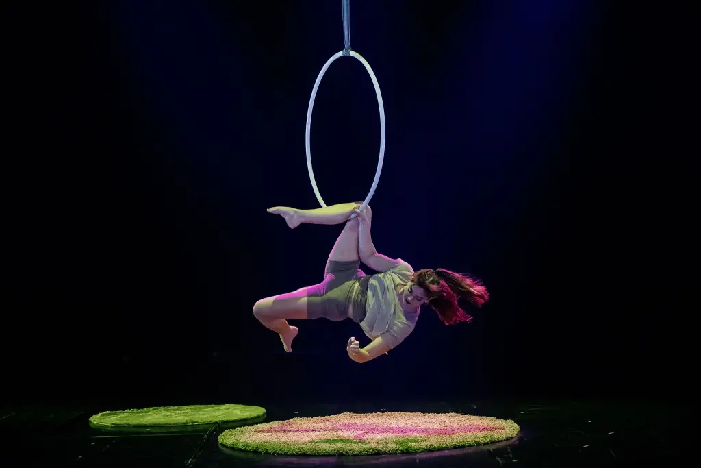 Une artiste de cirque, Kristina Shelton, est suspendue à un cerceau aérien, la tête en bas, sur une scène sombre, éclairée par des lumières violettes et vertes, avec deux tapis circulaires en dessous.