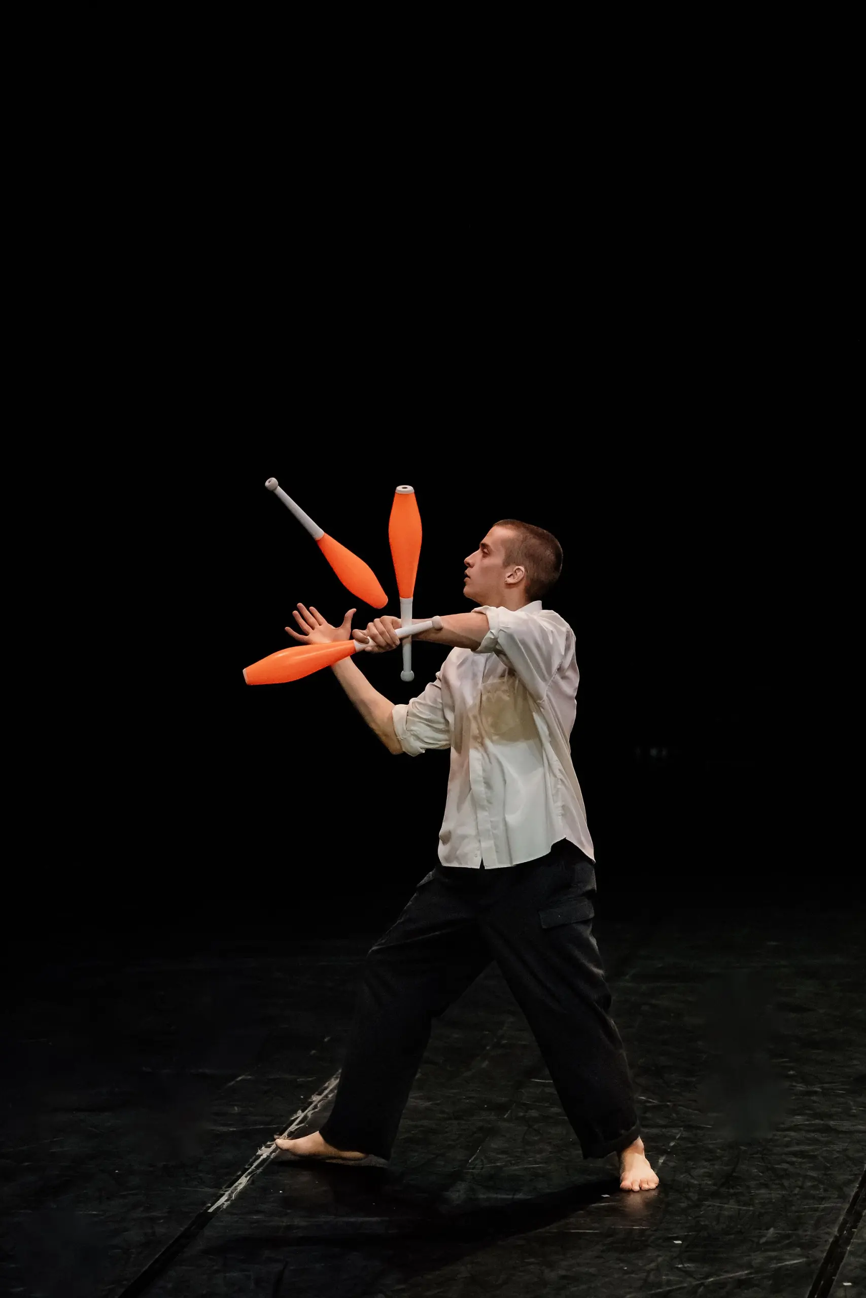 Nadav Sadlik, pieds nus, vêtu d'une chemise blanche et d'un pantalon noir, jongle avec quatre massues orange sur une scène sombre au fond noir.