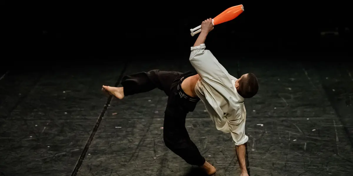 Nadav Sadlik, vêtu d'une chemise blanche et d'un pantalon noir, se tient en équilibre sur une main tout en tenant des massues de jonglage, l'une d'entre elles étant posée sur le sol, sur une scène sombre.