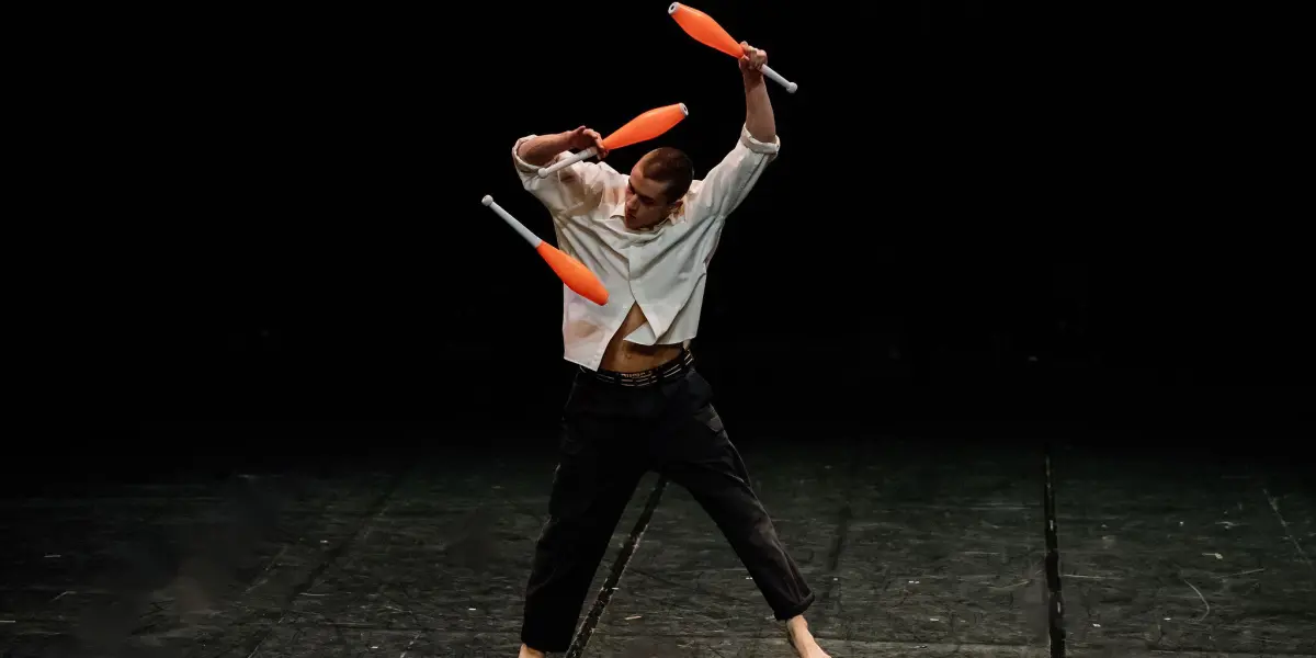 Nadav Sadlik, un artiste pieds nus, vêtu d'un pantalon noir et d'une chemise blanche, jongle avec trois massues orange sur une scène sombre, prenant une pose dynamique avec les bras levés et les jambes écartées.