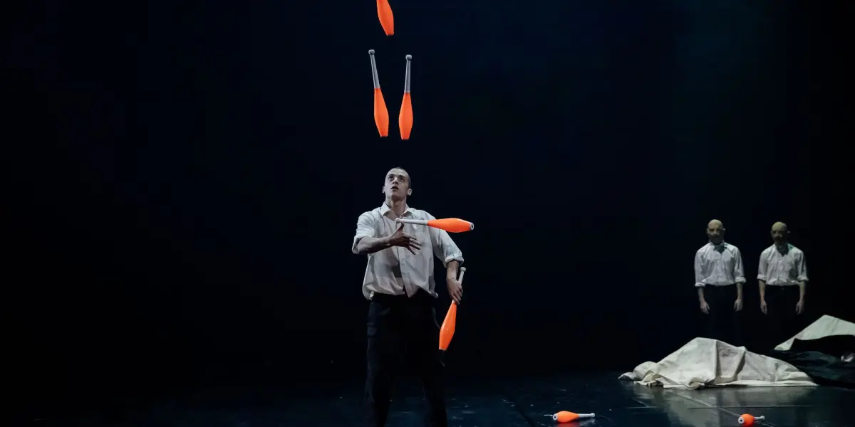 Un artiste pieds nus, peut-être Nadav Sadlik, vêtu d'une chemise blanche et d'un pantalon sombre, jongle avec cinq massues orange sur une scène sombre, tandis que deux personnes en tenue similaire se tiennent à l'arrière-plan, près d'un drap blanc.