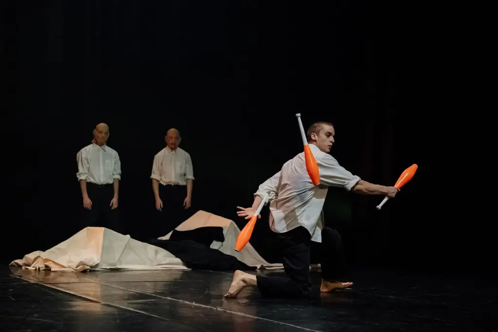 Un artiste pieds nus, Nadav Sadlik, porte des massues de jonglage orange sur ses bras et ses épaules, tandis que deux personnes vêtues de chemises blanches et de masques se tiennent à l'arrière-plan, près d'un tissu drapé sur une scène sombre.