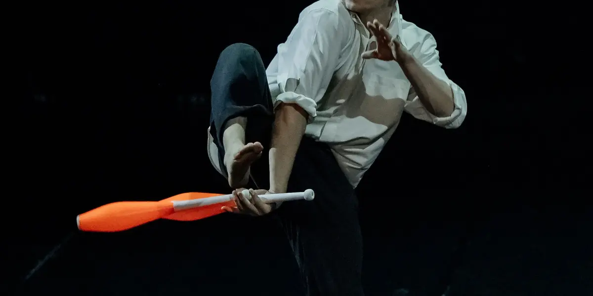 Nadav Sadlik, pieds nus, vêtu d'une chemise blanche et d'un pantalon noir, jongle avec des massues orange sur une scène sombre - il fait tenir une massue en équilibre sur son pied levé tandis qu'une autre tourne gracieusement dans les airs.