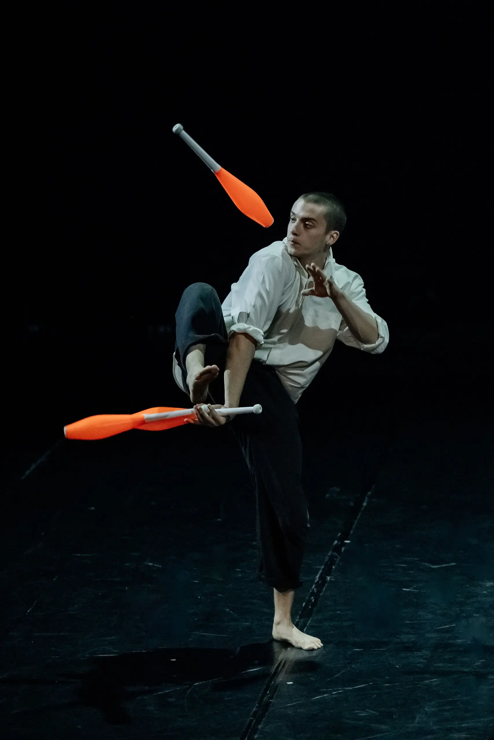 Nadav Sadlik, pieds nus, vêtu d'une chemise blanche et d'un pantalon noir, jongle avec des massues orange sur une scène sombre - il fait tenir une massue en équilibre sur son pied levé tandis qu'une autre tourne gracieusement dans les airs.