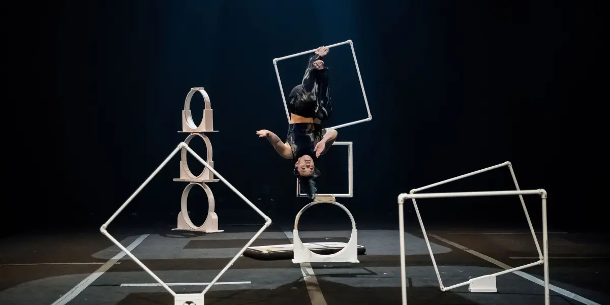 Un artiste vêtu de vêtements sombres, peut-être Haley Tenn, prend une pose acrobatique, suspendu à l'envers à l'intérieur d'un grand cadre carré sur la scène. Des formes géométriques et des structures sont disposées autour d'eux sous un éclairage dramatique.